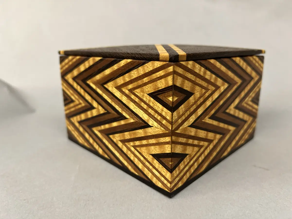 A hexagonal wooden box with intricate geometric inlay patterns of alternating dark and light wood, featuring concentric…