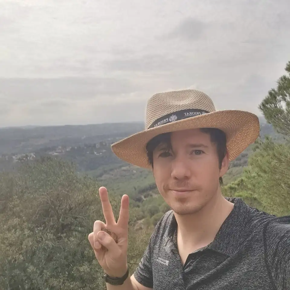 A man wearing a straw hat with a 'PRIORAT' band and a dark patterned polo shirt poses for a selfie outdoors, making a peace…