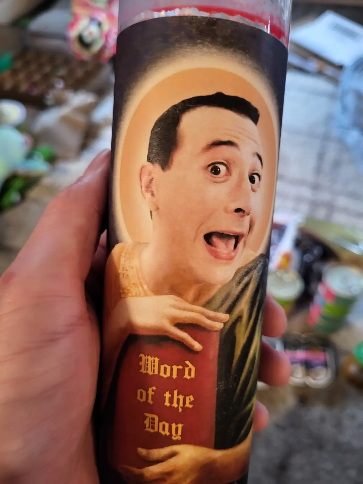 A hand holds a prayer candle featuring a saintly portrait of a man with wide eyes and an open mouth, holding a book labeled…