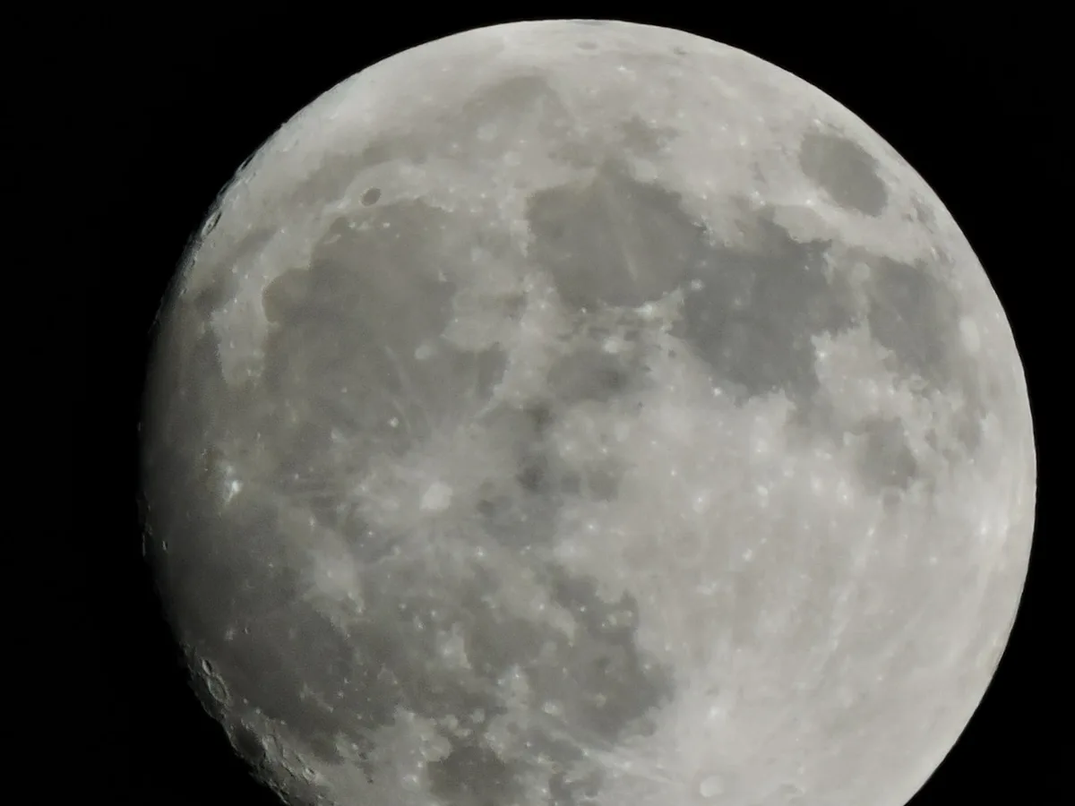 A close-up photograph of the moon against a black sky, showing its cratered, gray surface with visible maria and highlands…