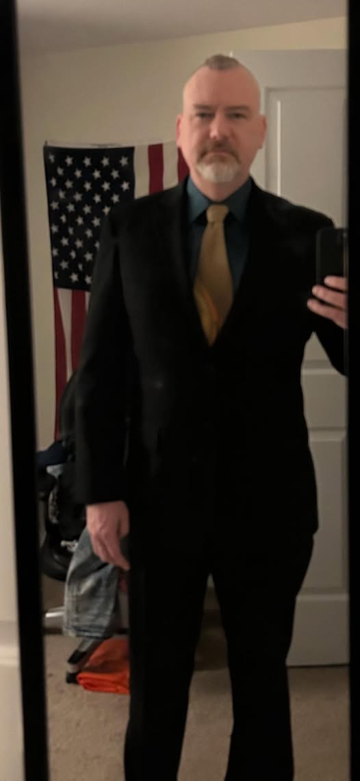 A man in a black suit and gold tie stands in front of a mirror, holding a phone to take a selfie. Behind him, an American…