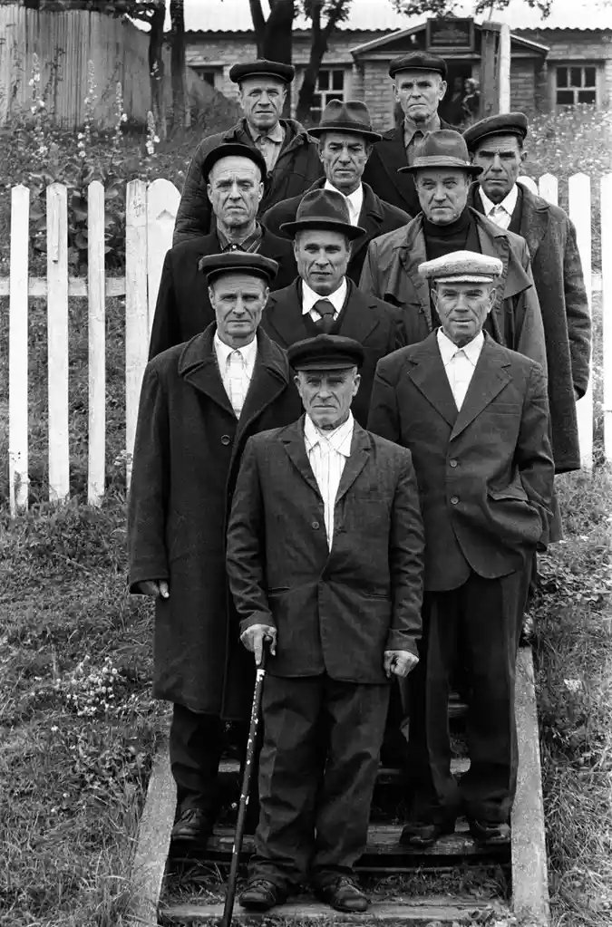 A black-and-white photograph shows a group of ten elderly men posing formally outdoors on wooden steps, wearing coats, hats…