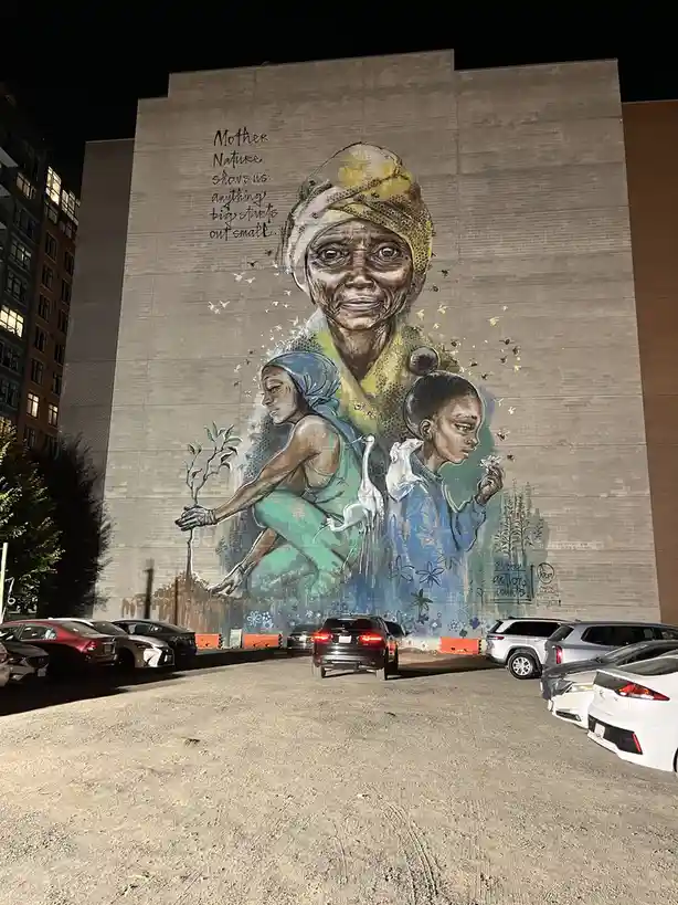 A large nighttime mural on a brick building depicts an elderly woman with a yellow headwrap, surrounded by bees and smaller…