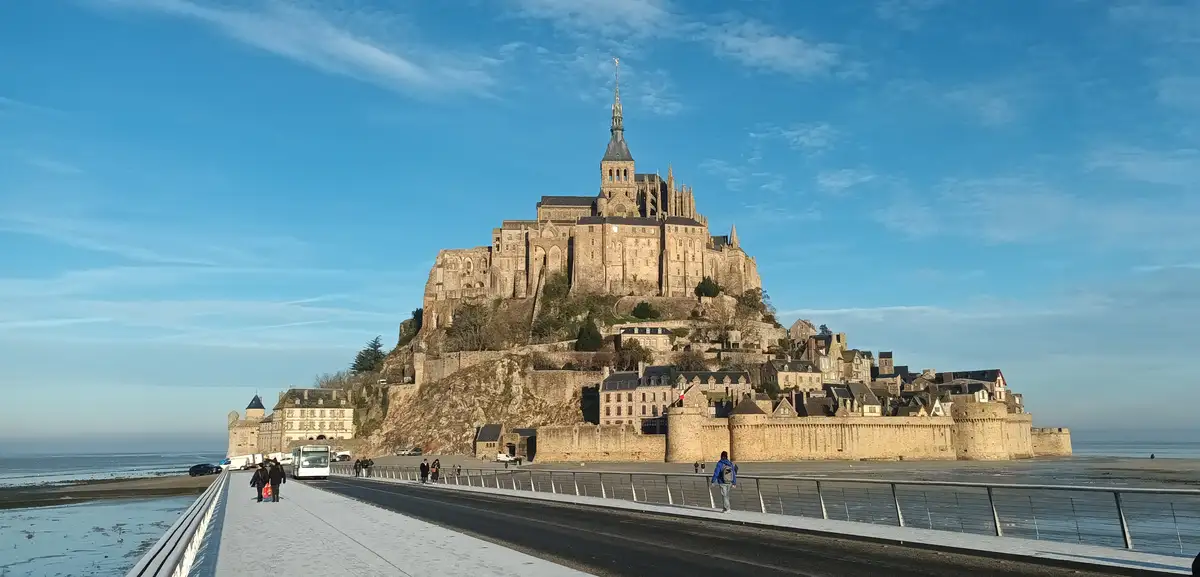 A wide-angle photo shows Mont Saint-Michel, a historic abbey on a tidal island in France, viewed from a modern causeway…