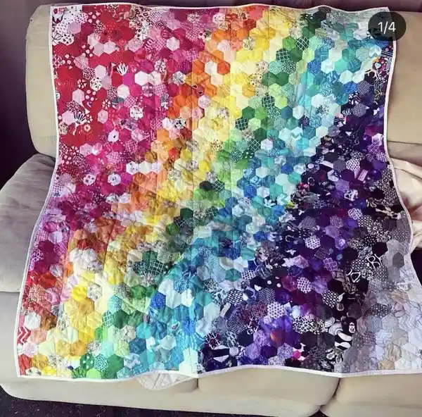 A colorful quilt made of hexagonal fabric patches arranged in a rainbow gradient from red to purple, draped over a beige…