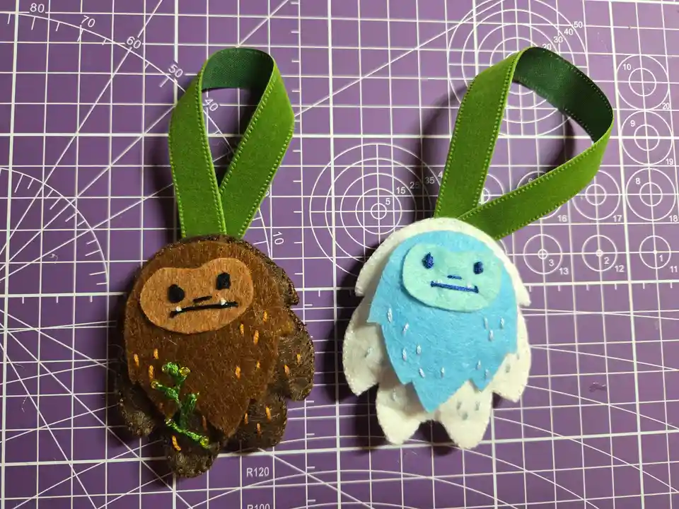 Two handmade felt ornaments, one brown resembling a grumpy forest creature and one blue and white resembling a snow monster…