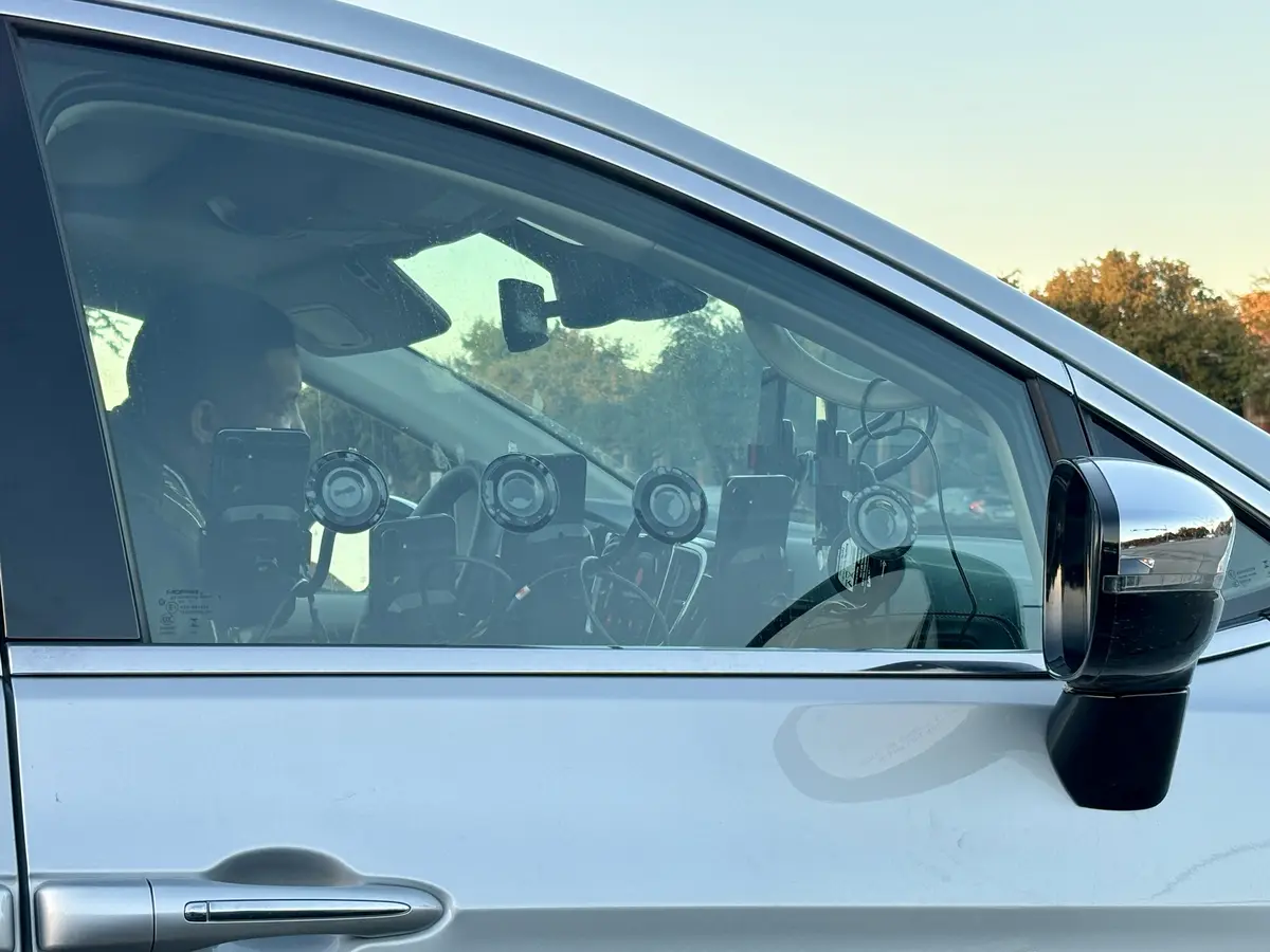 A silver car's passenger side window shows a person inside, surrounded by multiple phone mounts and recording equipment. The…