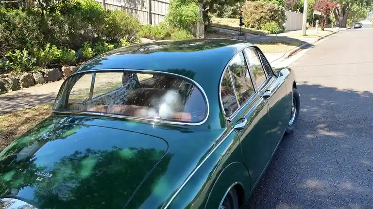 A glossy green classic Jaguar 3.4 is parked on a suburban street, its shiny body reflecting trees and sky. The overlaid text…
