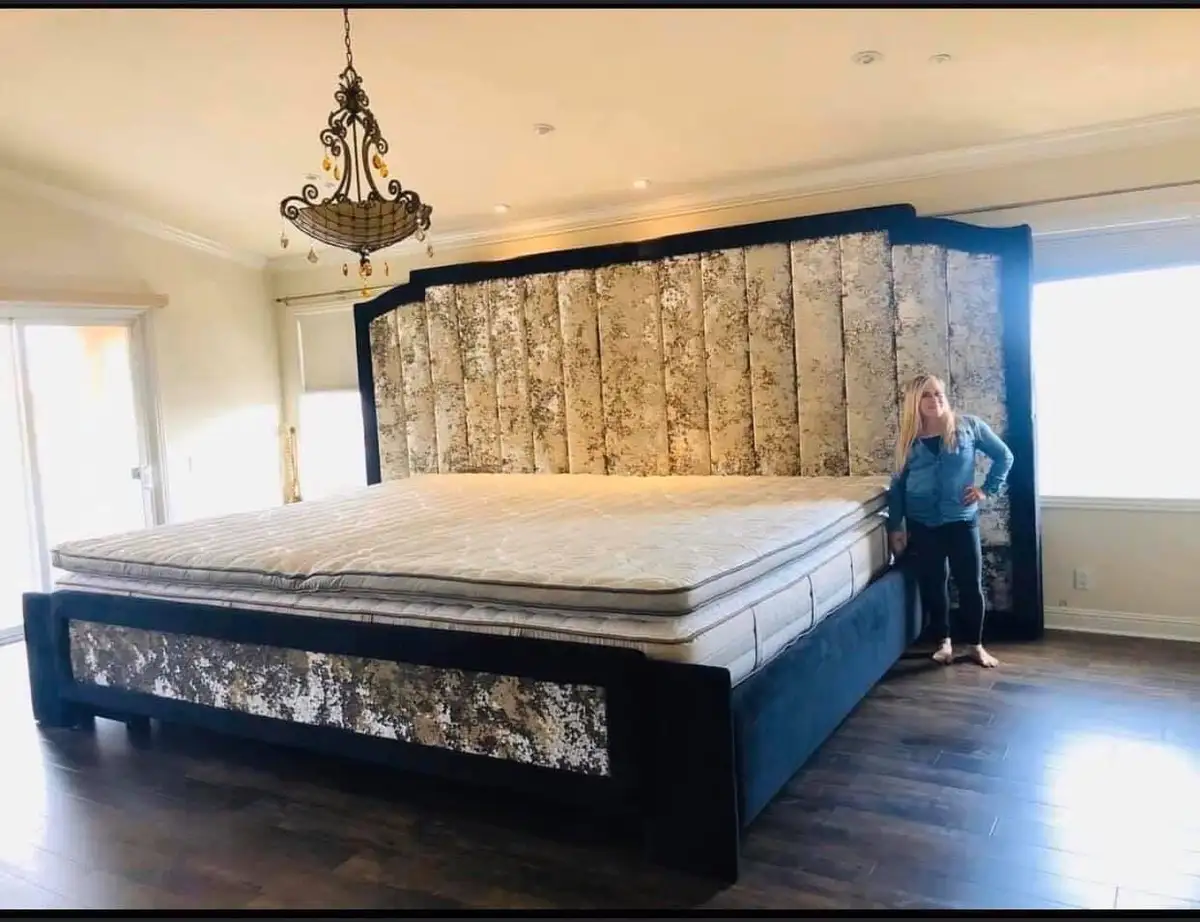 a woman stands beside an enormous bed with a tall, ornate headboard and a thick mattress, looking small in comparison. The…