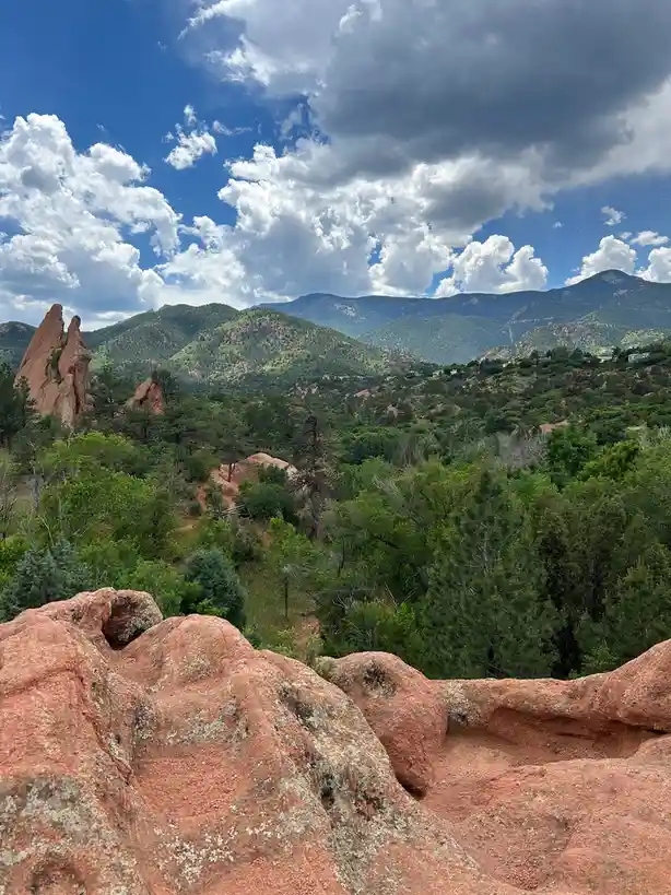 A scenic photo of red rock formations in the foreground with green forests and distant mountains under a partly cloudy blue…