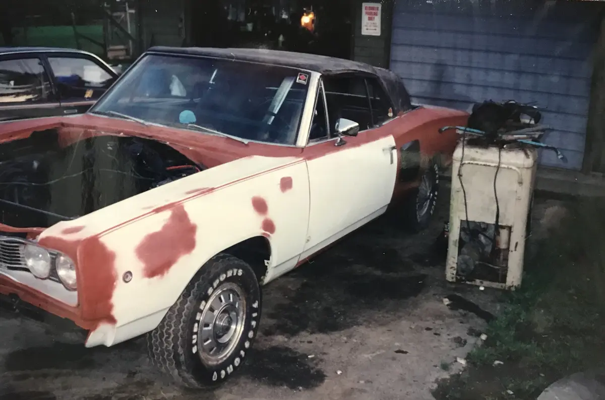 A red-and-white classic American muscle car with its hood open and paint peeling, parked next to a broken refrigerator in a…