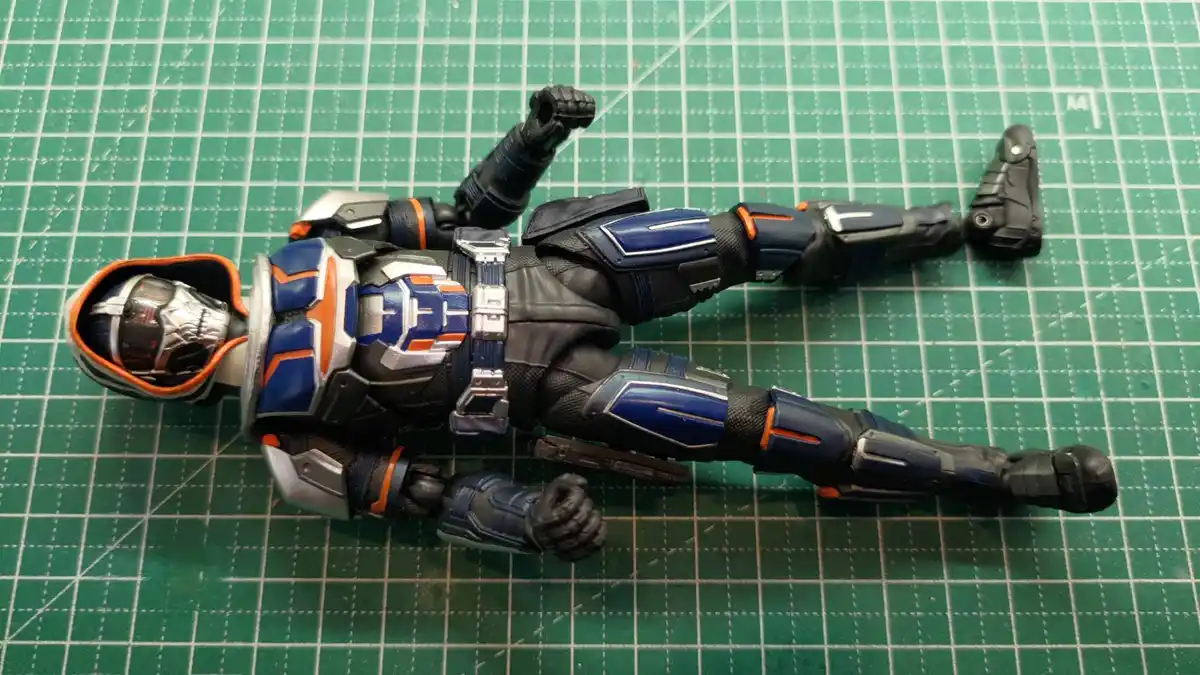 A blue, black, and orange armored action figure lies on its back on a green cutting mat, one foot detached and placed…