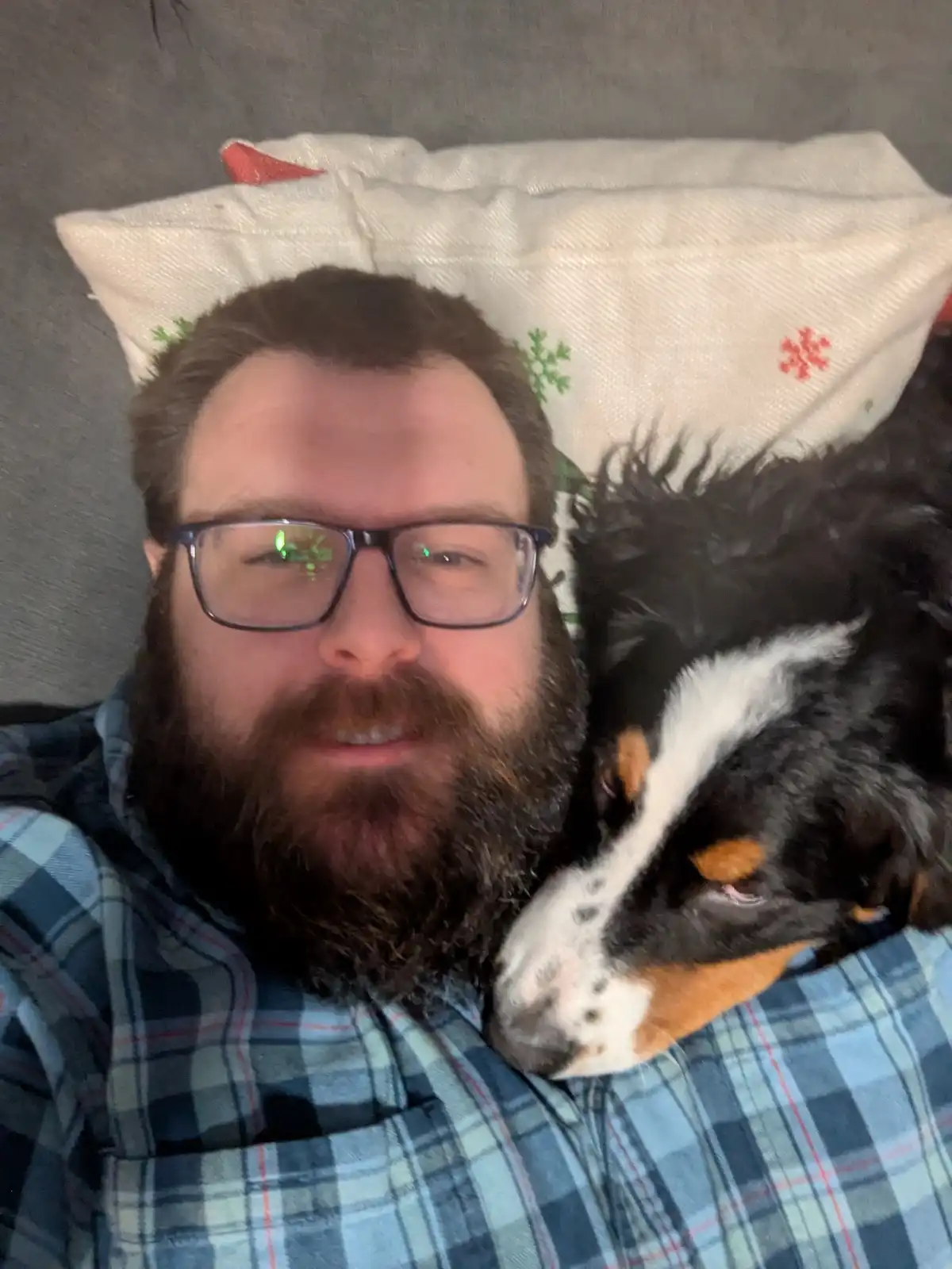 A man with a beard and glasses lies on a couch, wearing a plaid shirt, with a black, white, and tan dog resting its head on…