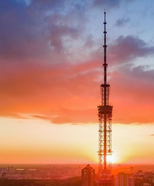 A tall, slender telecommunications tower stands silhouetted against a vibrant sunset sky with orange, pink, and purple…