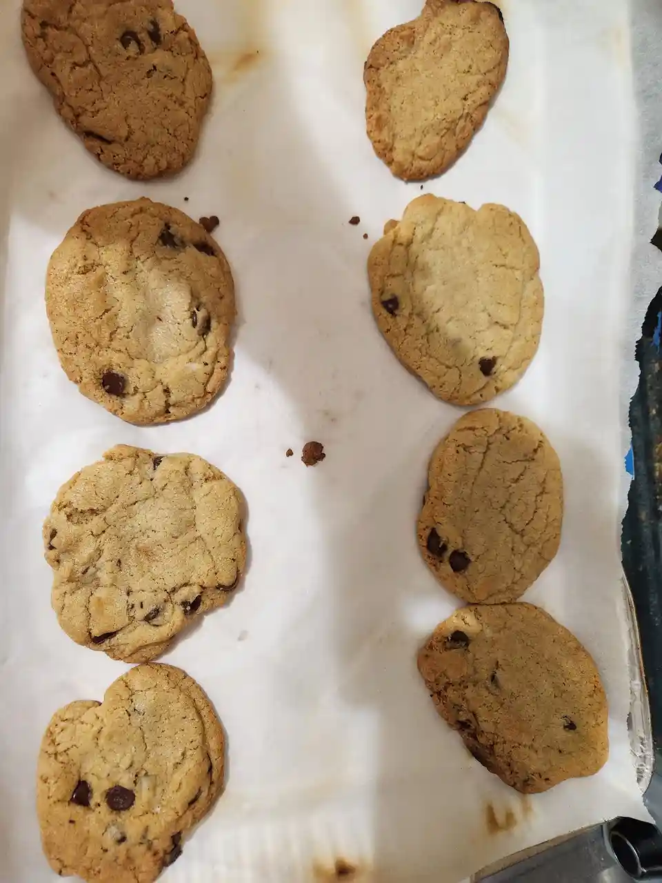 A baking tray lined with parchment paper holds eight chocolate chip cookies, unevenly baked with varying sizes and shapes…
