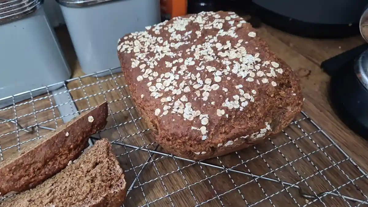 A loaf of dark brown bread topped with rolled oats sits on a metal cooling rack, with two slices cut and laid beside it on…