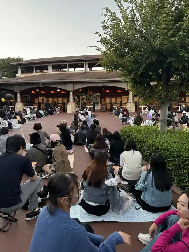 A large group of people are sitting on blankets and the ground in an outdoor plaza, facing a building with arches and…