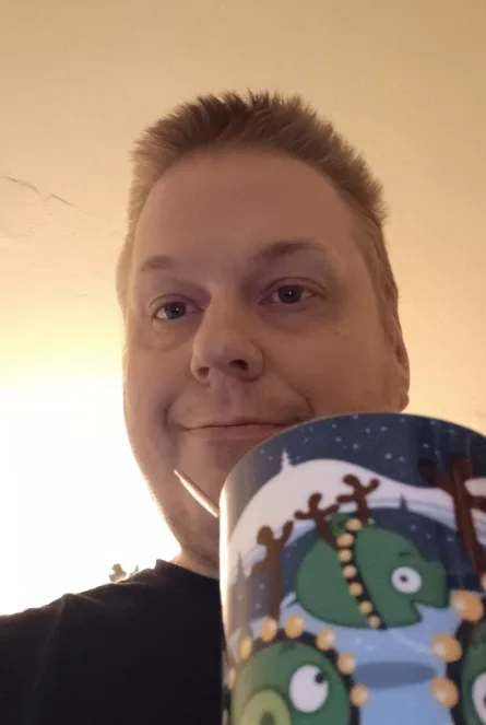 A man holds a mug featuring cartoon green pigs dressed as reindeer with antlers and beads, partially obscuring his face. He…