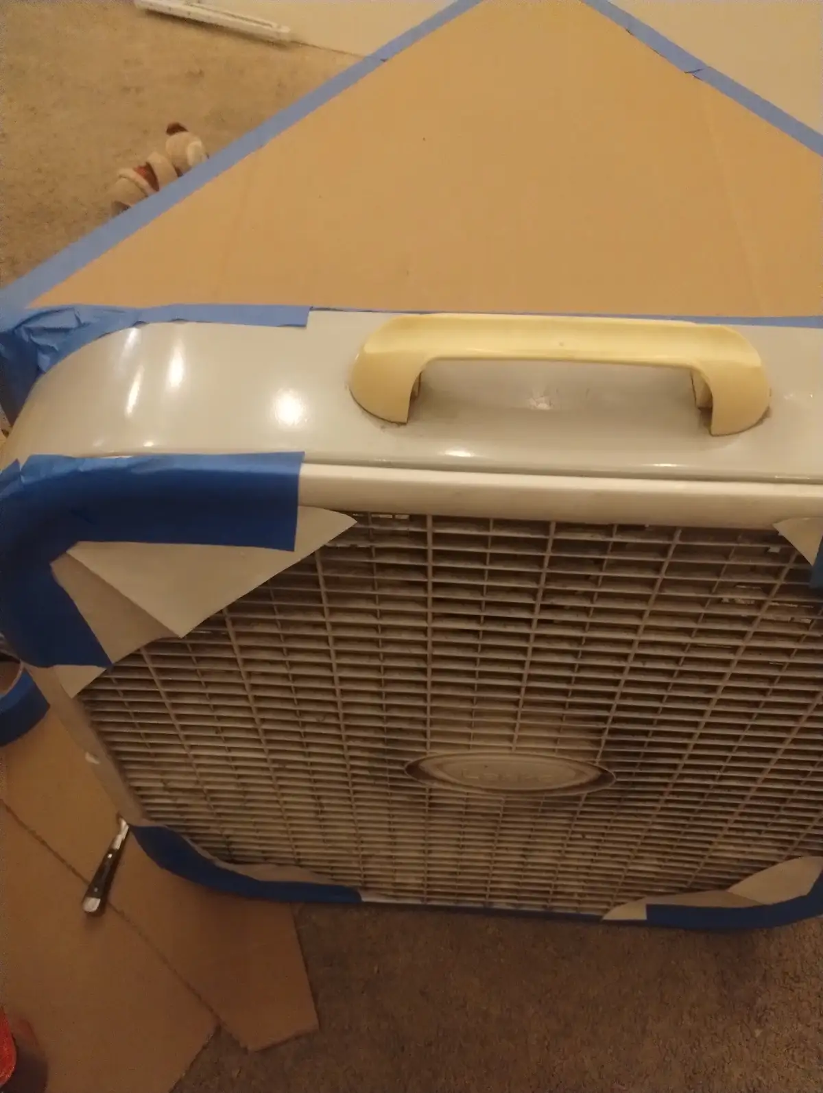 A dusty, beige portable air conditioner with a yellow handle is surrounded by blue painter's tape and cardboard, suggesting…