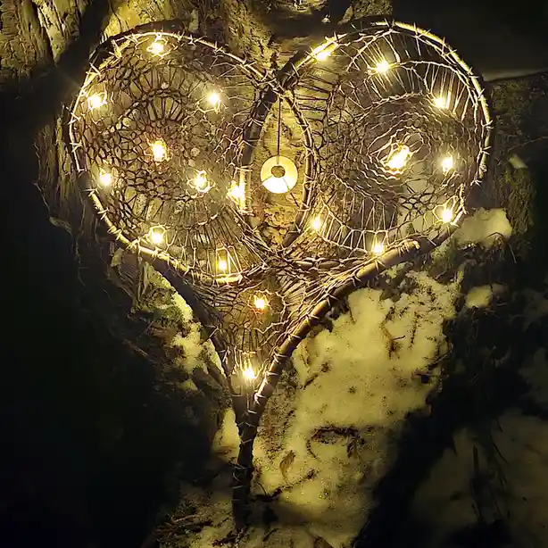 A heart-shaped decorative light fixture made of woven branches and glowing string lights is mounted on a tree in a snowy…