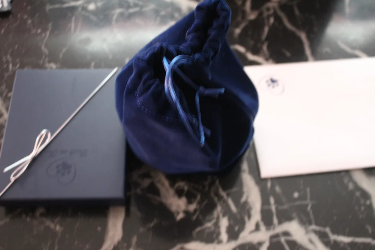 A dark blue velvet drawstring pouch sits on a black marble surface next to a dark blue box with a silver ribbon and a white…