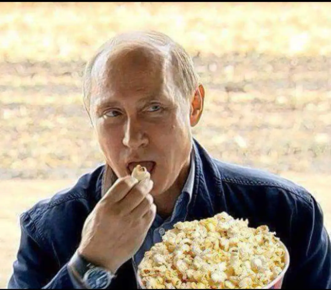 a man with a receding hairline wearing a dark jacket and watch is eating popcorn from a large red bucket while looking off…