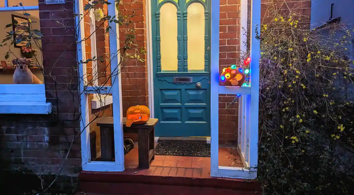 A nighttime photo of a house entrance with a teal door, brick walls, and Halloween decorations. Two carved pumpkins are…