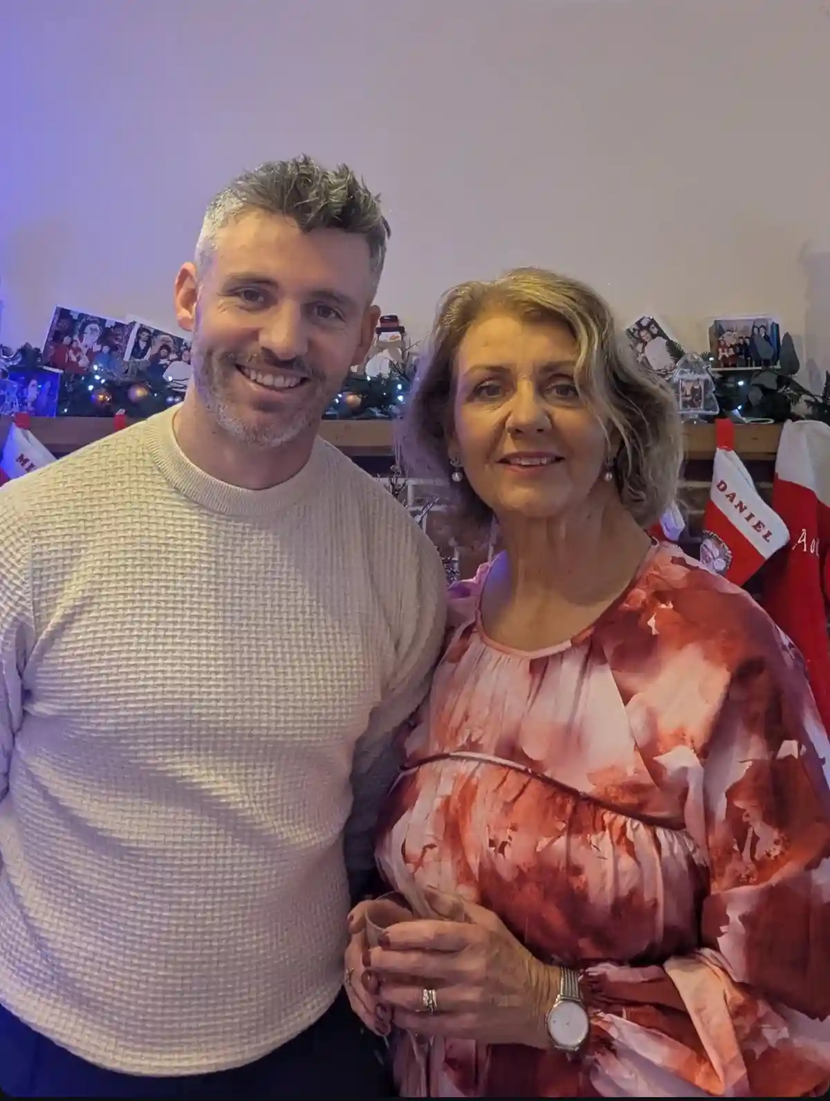 A man and a woman stand together in a Christmas-decorated room, smiling for the camera. The woman, wearing a pink and red…