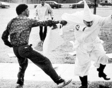 A black-and-white photo shows a man in a plaid shirt and dark pants kicking a man dressed in a white Ku Klux Klan robe and…
