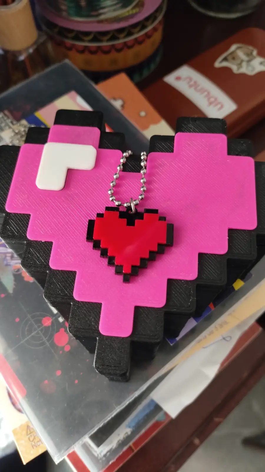 A stack of 3D-printed pixel art cards shaped like a pink and black character, with a red pixel heart pendant on a silver…