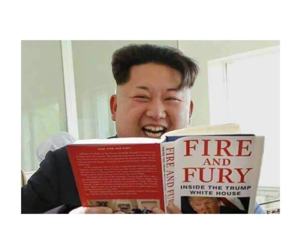 A man with a distinctive hairstyle and wide, toothy grin is holding open a book titled 'FIRE AND FURY INSIDE THE TRUMP WHITE…