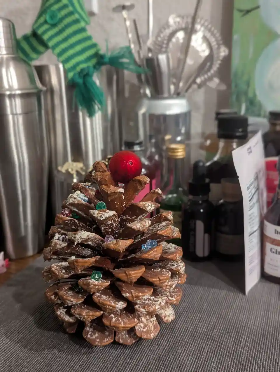A large pinecone decorated with white powder, colorful glass gems, and a red ornament sits on a gray countertop, surrounded…