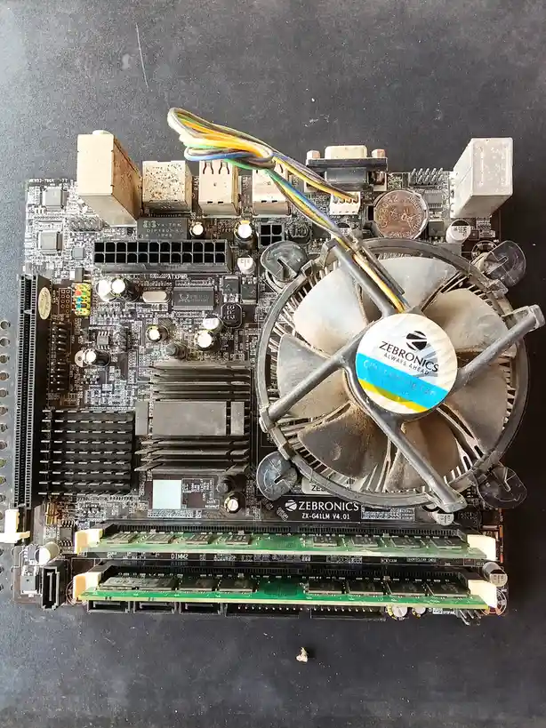 A dusty, old computer motherboard with a ZEBRONICS CPU cooling fan mounted on it, showing visible wear and dirt. The fan's…