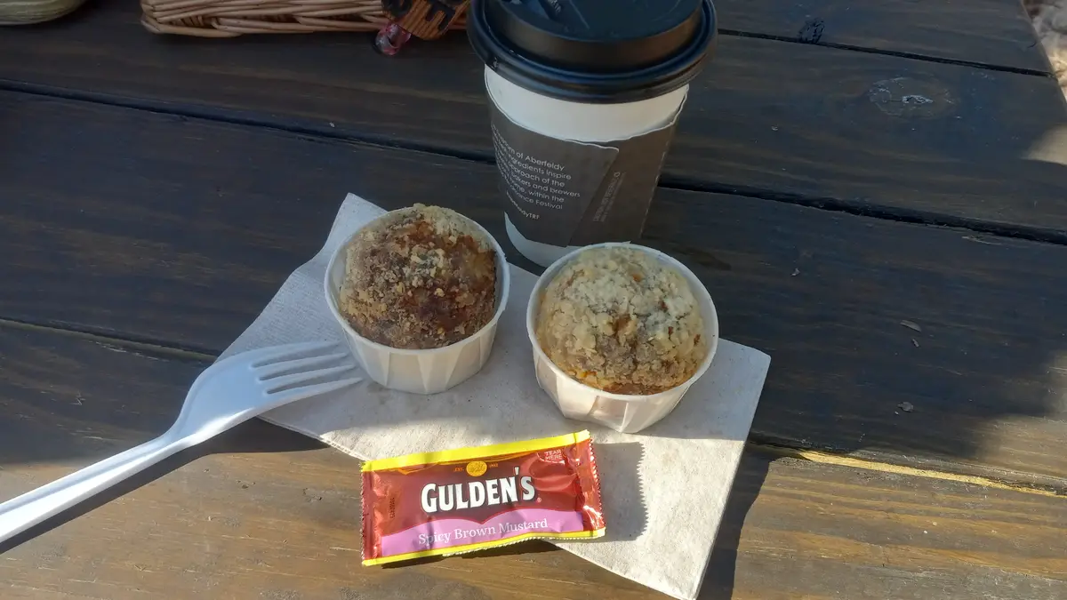Two muffins in white paper cups sit on a napkin next to a coffee cup and a plastic fork on a wooden table. A packet of…