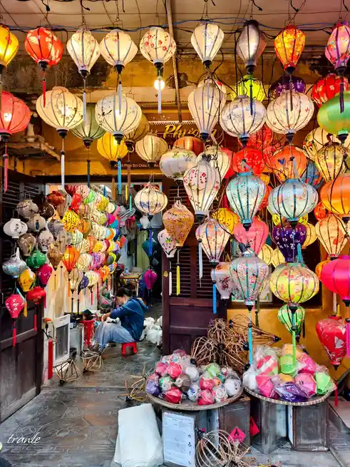 A vibrant market stall is densely packed with hundreds of colorful, illuminated paper lanterns of various shapes and…