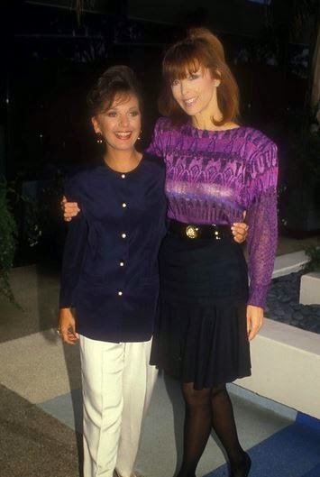 Two women pose together in a photo; one wears a navy blue button-up shirt and white pants, the other wears a purple…