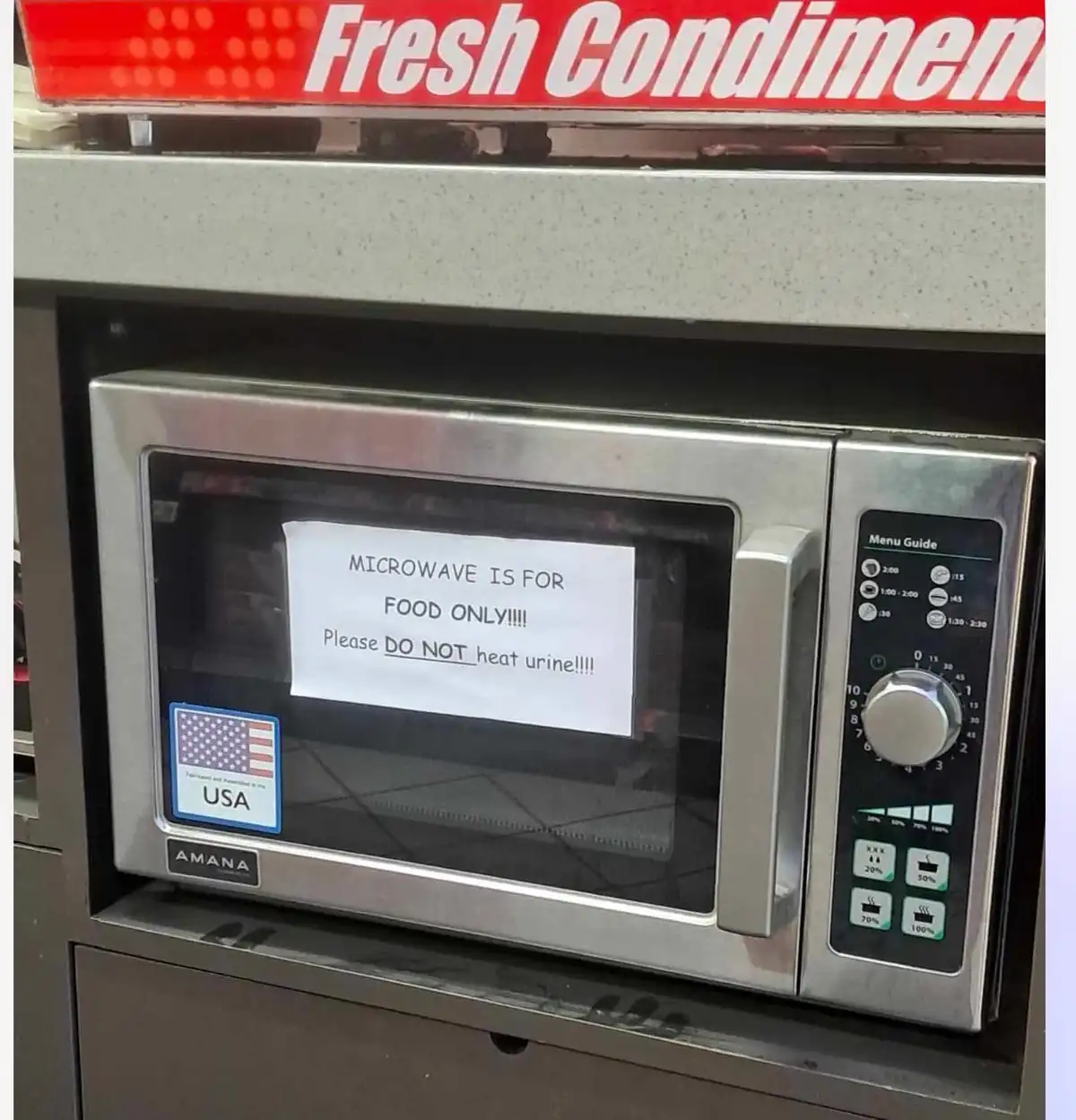 A stainless steel Amana microwave sits in a commercial kitchen under a 'Fresh Condiments' sign. A handwritten note taped to…