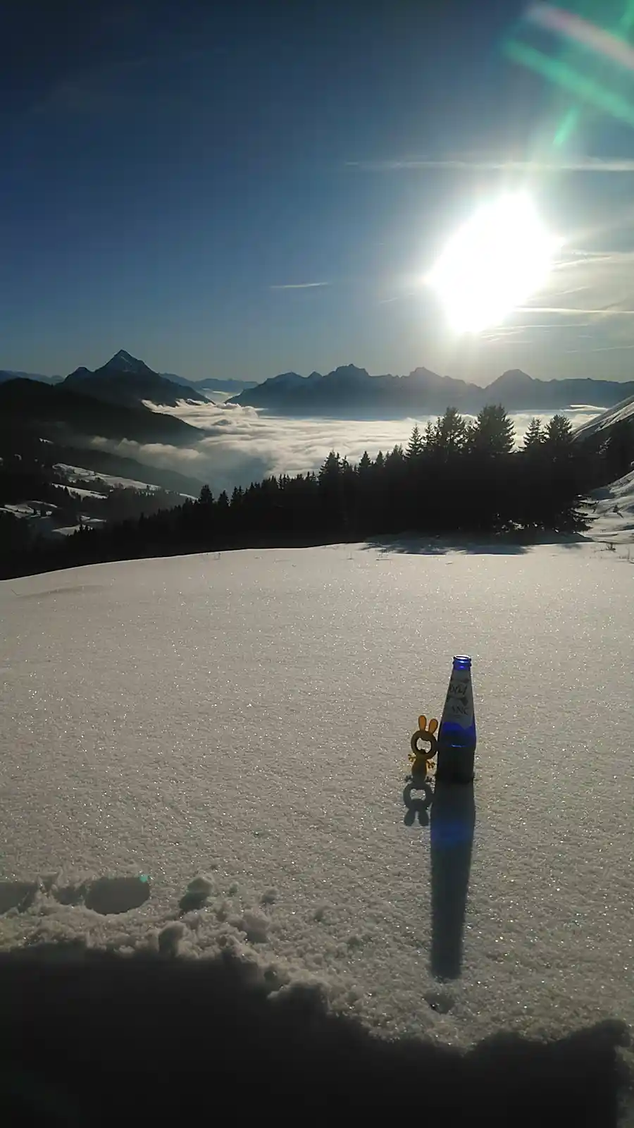 A blue glass bottle of 604 Blanc beer and a small yellow figurine stand upright in fresh snow on a mountain ridge. Behind…