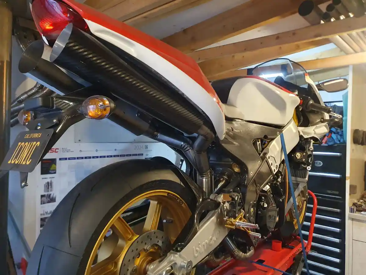 A red, white, and black sport motorcycle with carbon fiber bodywork and gold wheels is shown in a garage, mounted on a red…