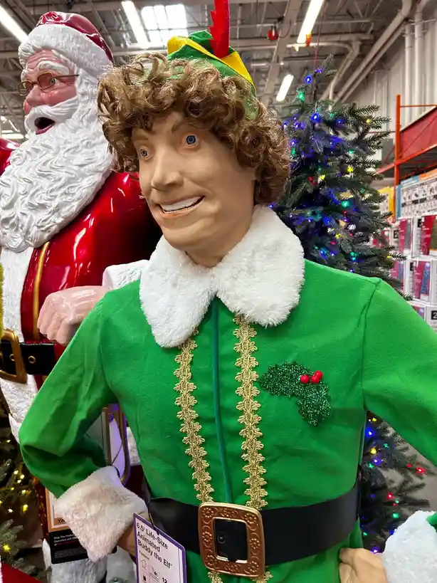 A life-sized animated Buddy the Elf mannequin in a green elf costume with a white fur collar and a holly pin, standing in a…