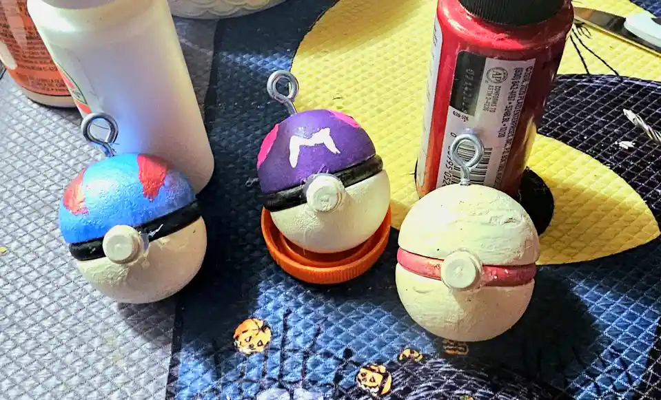Three handmade, crudely painted Pokémon Poké Ball crafts sit on a patterned tablecloth next to craft supplies like paint…