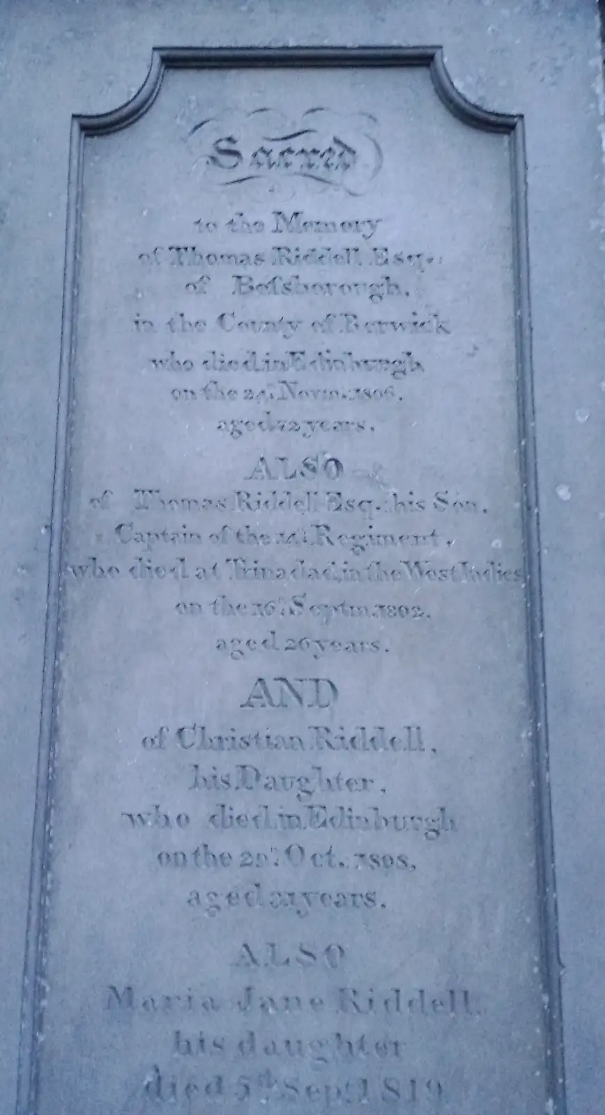 A close-up of an old, weathered grey stone memorial plaque with engraved text listing the deaths of four members of the…