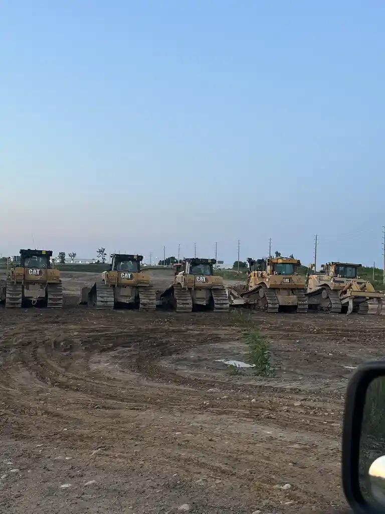 Five yellow Caterpillar bulldozers are parked in a row on a muddy construction site under a clear blue sky. The image is…