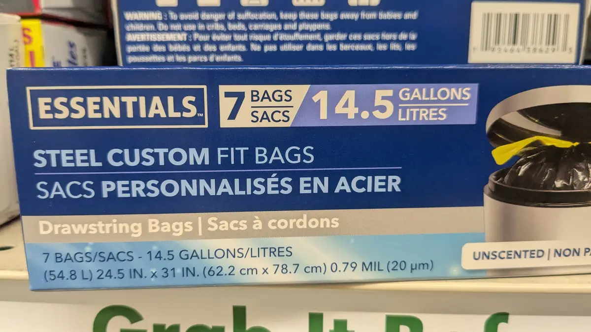 A close-up of a blue box of 'Essentials' brand steel custom fit drawstring trash bags, showing text that says '7 BAGS / 14.5…