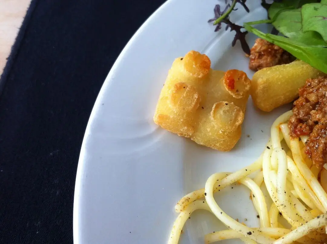 A close-up photo of a white plate with spaghetti, meat sauce, and a golden-brown fried food item shaped exactly like a Lego…