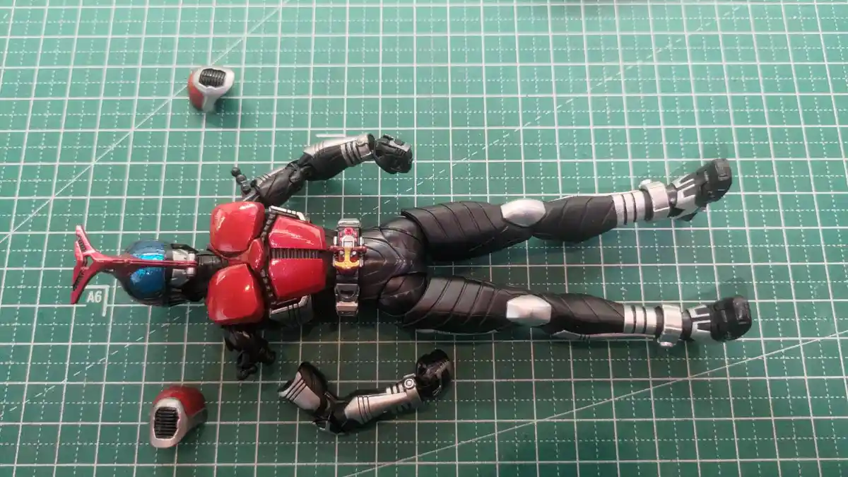 A red and black Kamen Rider action figure lies on its back on a green cutting mat, with its head, arms, and legs detached…