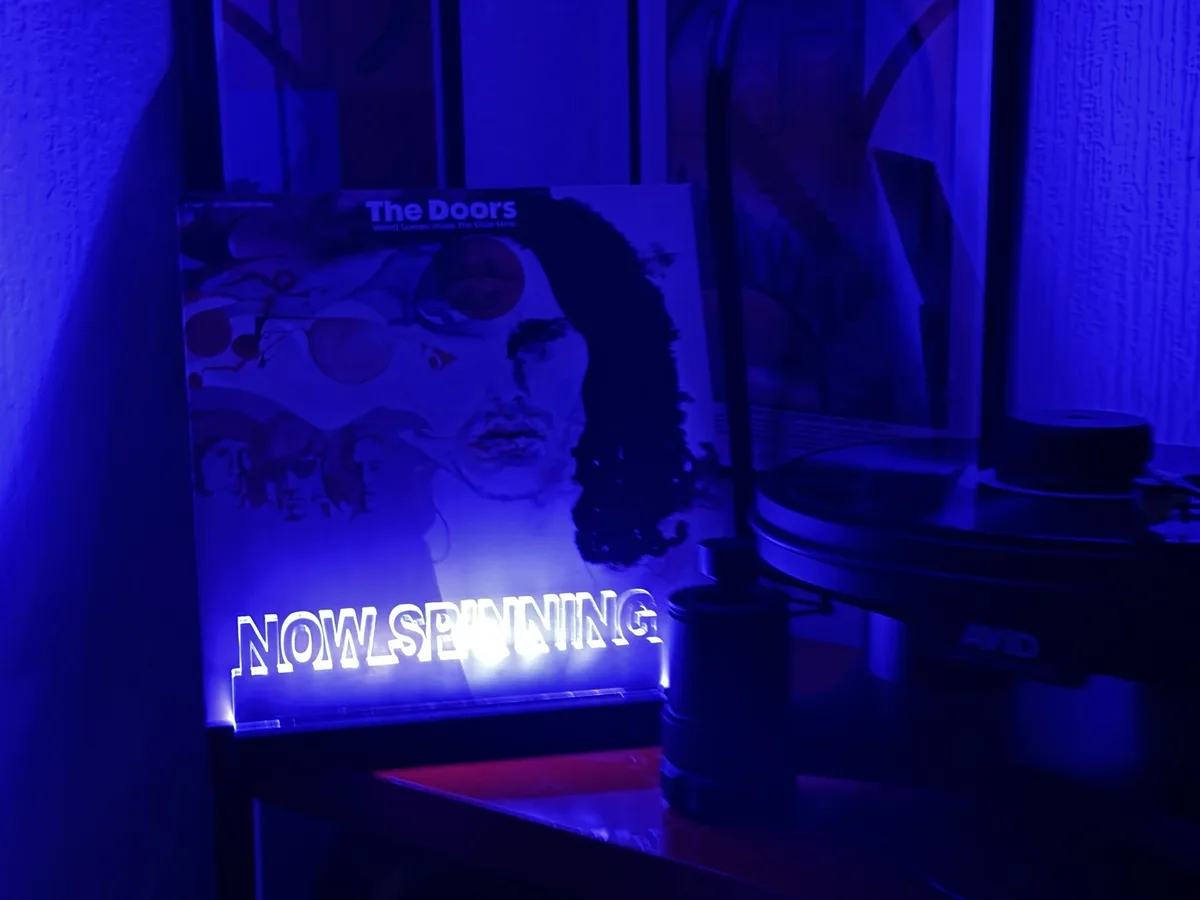 A vinyl record player sits under blue light with a 'The Doors' album cover leaning against it. A glowing sign in front of…