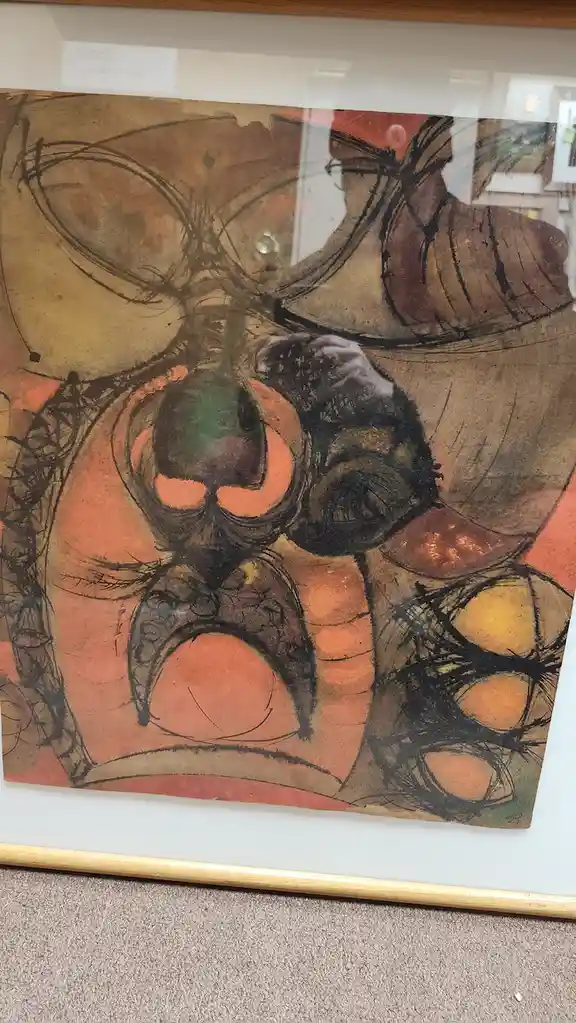 A framed abstract painting with swirling, organic shapes in earthy tones of brown, orange, and black, displayed behind…