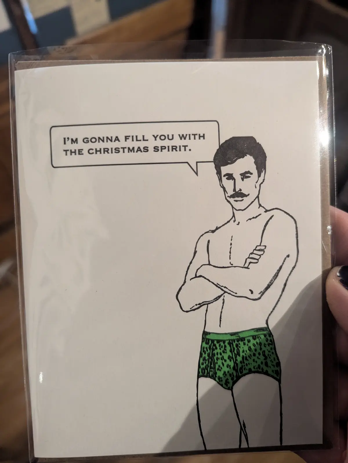 A hand holds a greeting card featuring a black-and-white line drawing of a shirtless man with a mustache and arms crossed…