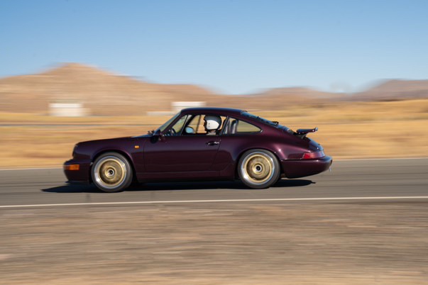 A maroon Porsche 911 is speeding down a desert road, with blurred background hills and motion blur on the wheels. The driver…